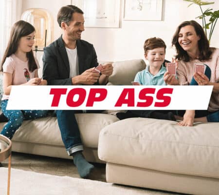 Familie spielt im Wohnzimmer auf dem Sofa einTOP ASS Kartenspiel von ASS Altenburger zusammen. Darüber liegt das TOP ASS Logo.