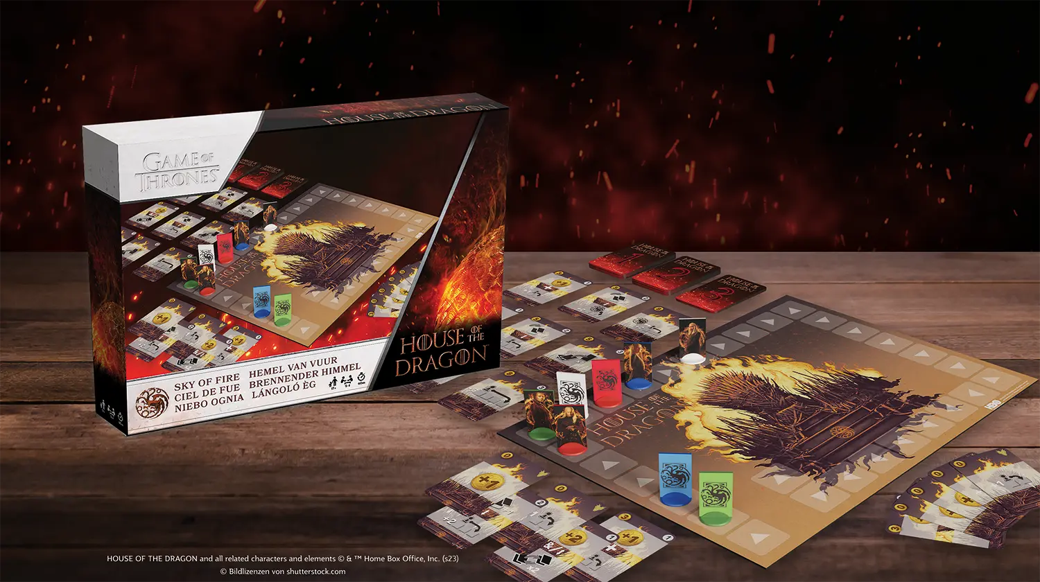 Es sind die Spielkomponenten des Game of Thrones House of the Dragon Sky of Fire Spiels auf einem Holztisch zu sehen, im Hintergrund steht die Schachtel.