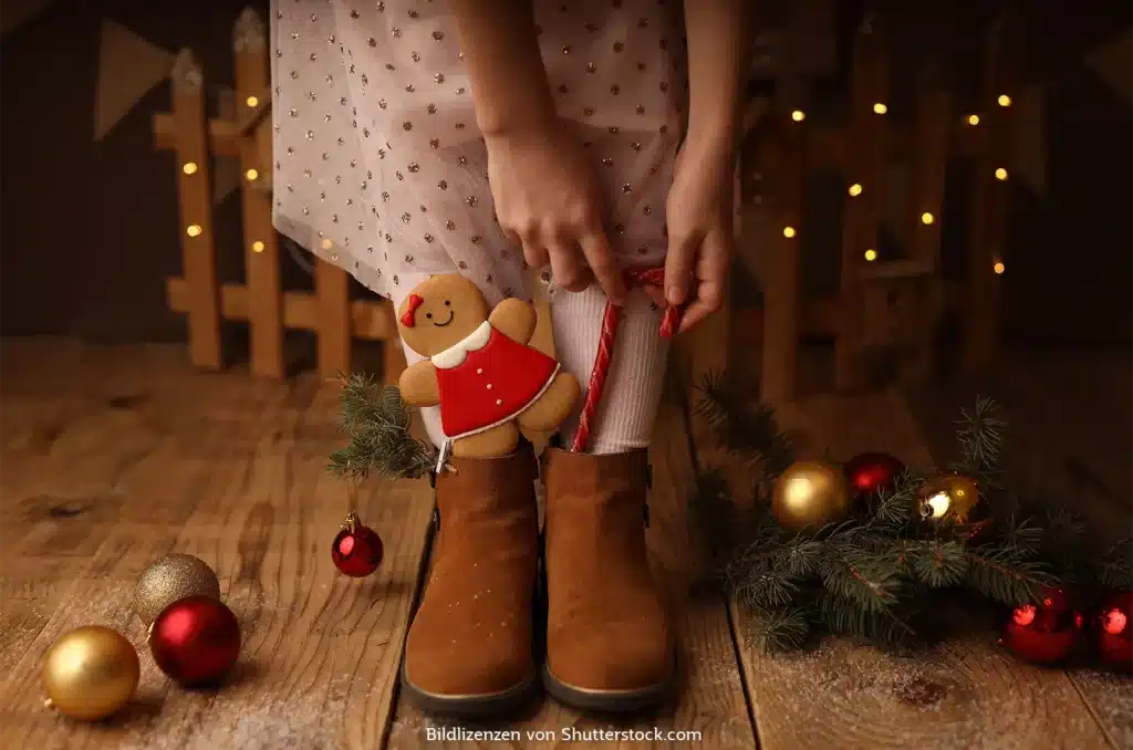 Beine und Füße eines Mädchens in braunen Stiefeln umringt von Weihnachtsdeko. Ein Lebkuchen und eine Zuckerstange stecken auch darin. Kleine Geschenke zu Weihnachten, ASS Altenburger